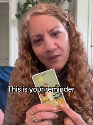 A woman with long curly brown hair is centered in the frame, holding a tarot card and speaking directly to the camera. The background is slightly blurred, featuring hints of a window, plants, and a blue blanket. The text overlay 'This is your reminder' is at the bottom.
