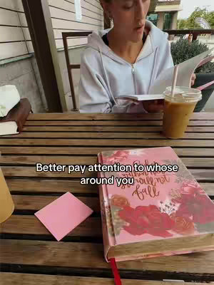 A woman is sitting at a wooden table on an outdoor patio, reading a book and sipping an iced coffee. In the foreground, there's a floral-covered journal, while a small pink sticky note rests on the table next to it. A white wall and greenery are visible in the background.