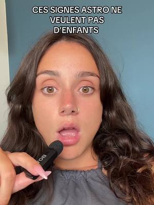 A young woman with brown hair and a nose ring is looking at the camera with her mouth open, holding a microphone. Text overlayed on the image reads "CES SIGNES ASTRO NE VEULENT PAS D'ENFANTS". The background is a plain blue wall.