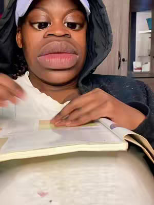A close-up shot features a person with a hooded jacket, a white t-shirt, and a white cap. A comical filter is applied, enlarging their lips and eyes. Their hands are holding and interacting with an open book on a table, creating a focused, but silly vibe, with a blurred background.