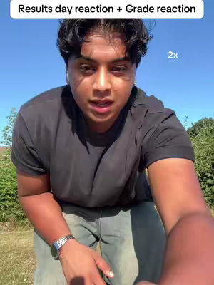 A person with dark hair and black shirt looking at the camera, shot from a low angle against a blue sky and green background.