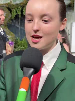 The cover image features Bella Ramsey, positioned mid-frame with the upper body visible. She wears a green suit with a white collared shirt and a red tie, holding a black microphone. A background of green foliage is visible. The shot is well-lit and appears to be taken at an event.