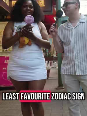 The video cover features two people in an outdoor setting. On the left, a woman in a white dress holds a small plush toy and a handheld fan, standing in front of a pink-toned backdrop. On the right, a man wearing glasses and a striped shirt holds a microphone, leaning in. The text overlayed in a red box reads 'LEAST FAVOURITE ZODIAC SIGN'.