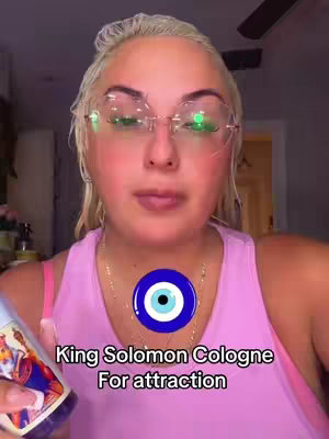 A selfie-style shot featuring a light-skinned woman with blonde hair wearing glasses and a pink tank top. Text overlays on the image promote "King Solomon Cologne" and its attraction properties. The woman is holding a bottle of cologne with an image of a king on it. A blue circular graphic is positioned in front of her chest.