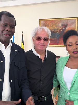 Three people posing for a photo. The background is an office setting, and the lighting is natural, with a painting and flag visible behind the subjects. A man on the left is wearing a black jacket over a white shirt, with a slight smile and a pose as if he's about to speak. In the center is a man wearing a black shirt and sunglasses, and on the right is a woman in a green blazer and a white shirt. The composition is a medium shot, focusing on the upper bodies and faces of the people, creating a formal and inviting feel.