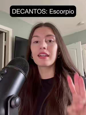 A woman with long brown hair is centered in the frame. Behind her, there's a microphone in the foreground on the left and a television screen in the background. The woman is gesturing with her hand. A text overlay with the words "DECANOS: Escorpio" in white text is positioned in a black rectangle on the top of the frame.