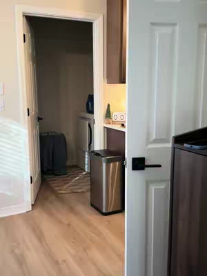 The image shows an interior shot of what appears to be a laundry room with a door slightly ajar, a shiny metal trash can in the middle of the frame, and a glimpse of another room. To the right, a white door with black hardware is visible. The floor is a light, wood-grain pattern. Overall, the scene is well-lit and suggests a clean, modern living space.