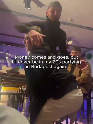 A photo taken at a night club in Budapest. A man in a black sweater is holding a woman in a party setting. A man is standing behind the woman dancing. Text at the bottom of the photo reading "Money comes and goes, but I'll never be in my 20s partying in Budapest again."