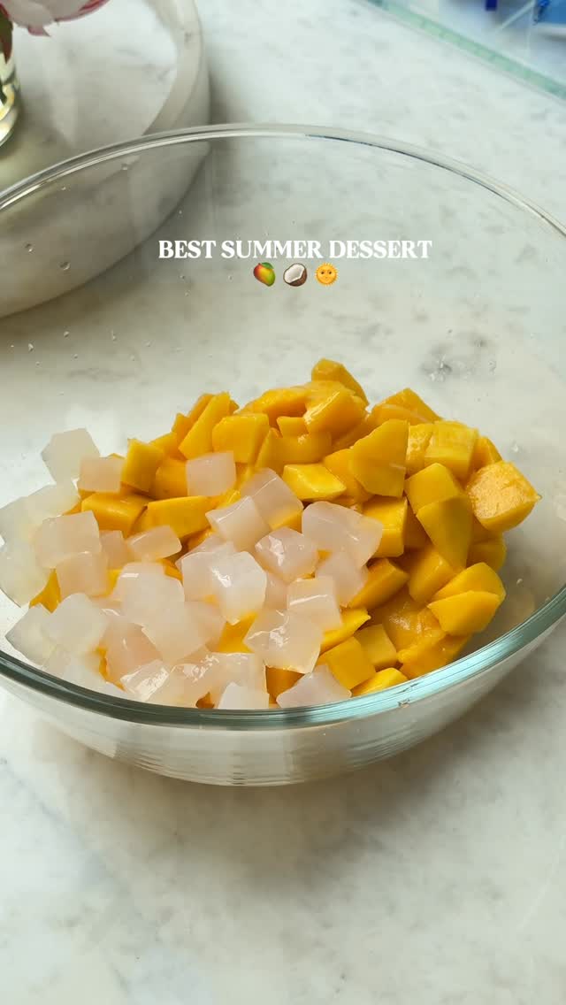 A clear glass bowl filled with diced yellow mango and white, cubed coconut jelly. The bowl sits on a white, marble-like surface. In the background, there's a glimpse of another container and a bit of the setting, likely a kitchen.
