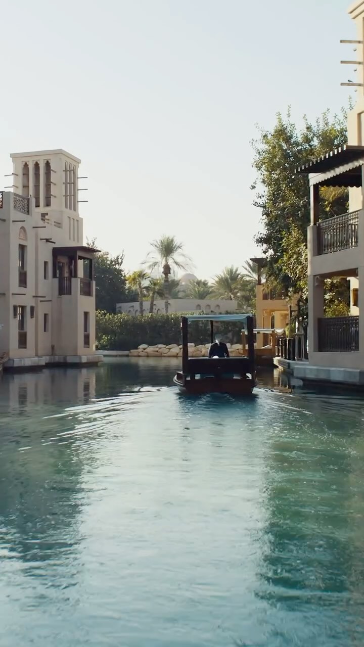 A serene, wide shot captures a tranquil waterway scene in Dubai. A small wooden boat glides through the turquoise water. Buildings with Middle Eastern architecture, featuring cream-colored walls, arches, and balconies line the water's edge. Palm trees and lush greenery are interspersed, and the bright sky creates a well-lit environment.