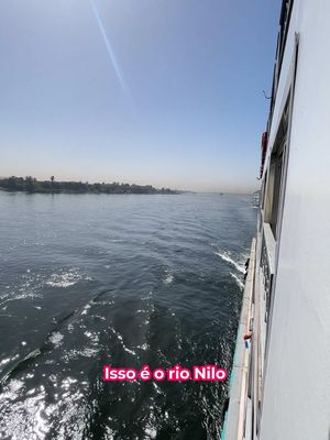 The image shows a view from the side of a boat, with the Nile River and sky visible. The right side of the image features the side of the boat, while the left side shows the river and a distant shoreline with trees. Above is a clear blue sky.