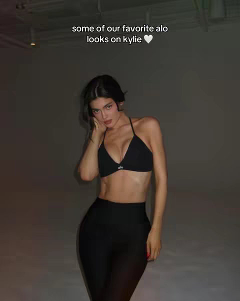 The image showcases a woman in a dark room, posing in athletic wear. The woman is a model, wearing a black sports bra and high-waisted black leggings. She has dark hair and is striking a pose, hand on her face, with a neutral backdrop.
