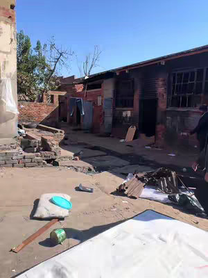 An outdoor scene of a dilapidated building with visible signs of destruction. A partially collapsed brick structure is on the left and a long, dark building with broken windows on the right. Piles of debris lie scattered on the ground, suggesting abandonment or recent demolition. The sky is clear, and the overall color palette is muted, reflecting a sense of decay.