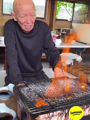 The image shows an older man with a bald head wearing a black long-sleeved shirt tending to a grill. The grill is filled with food cooking over flames. Nearby are small, dark-colored bowls and some other cooking tools on the counter. A lemon8 logo is in the corner. The background has windows and a portion of a white table behind them.