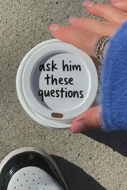 A close-up shot shows a woman's hand holding a coffee cup lid with the words "ask him these questions" written on it. The lid is white with a black font. The woman's fingers, painted with pink nail polish, are adorned with rings, and she wears a blue sleeve with a sweater texture in the background. The surface appears to be a cement ground, with visible sneakers.