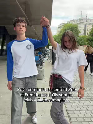 Two young people, one male and one female, holding hands with raised arms against an outdoor backdrop. The male on the left wears a blue and white shirt, and the female on the right is in a white shirt. The image is taken in an urban setting with a building and greenery visible.
