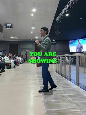 A man in a suit is speaking to an audience in a large hall. The image is framed with a vertical perspective, with the presenter on the left and most of the audience out of focus. In the foreground, a green text overlay reads "YOU ARE SHOWING."