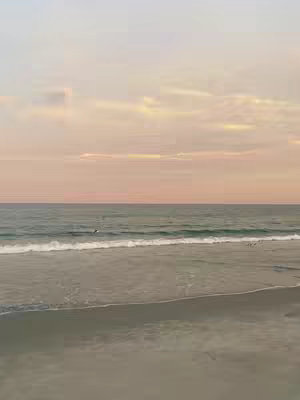 A serene beach scene at sunset. The image is divided horizontally with a light pink and orange sky taking up the top half, and the bottom half showing the ocean and the beach. The water has small waves with a possible person in the water, with a light sandy beach in the foreground. The overall effect is calm and peaceful.