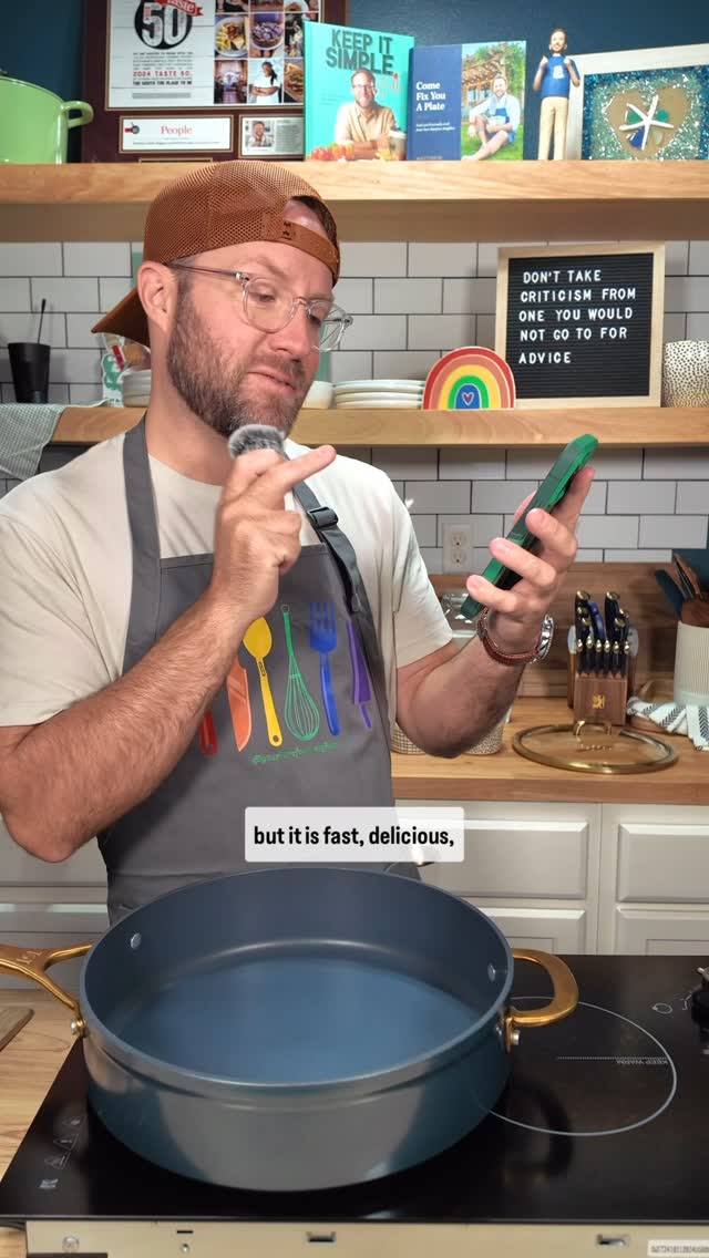 A man wearing glasses, a brown cap, and an apron stands in a kitchen, holding a microphone and a phone. Behind him are shelves with cookbooks, a sign, and decorative items. In front of him is a pan on a stove.