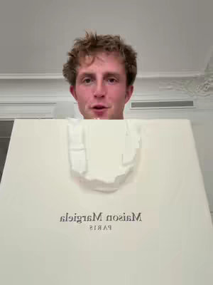 A person is holding up a white shopping bag with the text 'Maison Margiela PARIS' on it and is looking at the camera. The background appears to be a well-lit room.