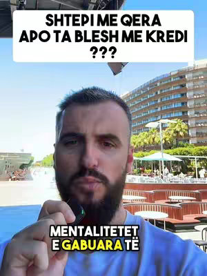 A man stands in front of a blurry outdoor background, holding a small microphone. Two text overlays are present: one above the man with a question about buying or renting property in Albanian, and another beneath with the words "MENTALITETET E GABUARA TË" in Albanian.