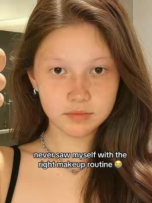 A selfie-style shot of a young person's face with brown hair, looking directly at the camera. The background appears to be a room with neutral lighting, placing the focus on the person. The person is wearing a black top and a silver chain necklace, and has subtle makeup. Overlaid text at the bottom reads, "never saw myself with the right makeup routine"