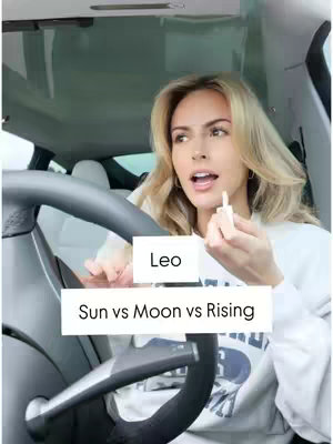 A selfie-style shot taken from inside a car.  A woman is in the driver's seat, partially obscured behind the steering wheel.  She has blonde hair and is holding a small item, possibly a makeup product.  The background shows the car's interior, with the windshield and side window visible.