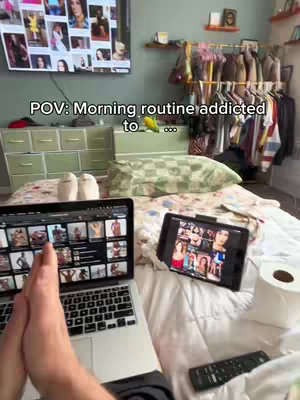 A bedroom scene with a person holding a laptop and a tablet in front of them while lying on a bed. The laptop displays images and videos of women, and the tablet shows the same content. A television is mounted on the wall, and a remote control and a roll of toilet paper can be seen on the bed. The room is lit naturally with a window out of frame.