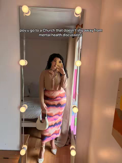 A woman in a mirror selfie, dressed stylishly in a neutral top and colorful skirt, holding a phone. Rows of lights frame the large mirror.