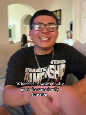 A man with glasses smiles widely, his face filling the frame. He wears a black t-shirt with white lettering and a silver chain. Text on the screen states, 'When THE cousins finally go to the same family function.' The background is a softly lit room with neutral tones.