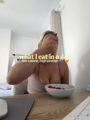A selfie-style shot of a woman partially covering her face with her hand while seated at a table. In front of her is a white bowl containing berries. The text overlay states: what I eat in a day, with the information about calorie and protein levels and a series of fruit/vegetable emojis.