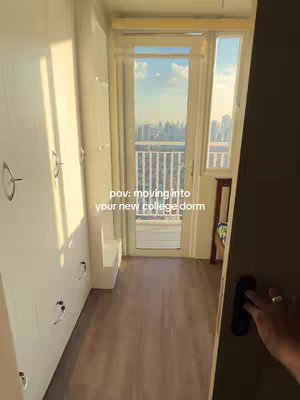 View from inside a room with a door and window facing outdoors. The interior has light-colored walls and wooden floors. A hand opens the door.