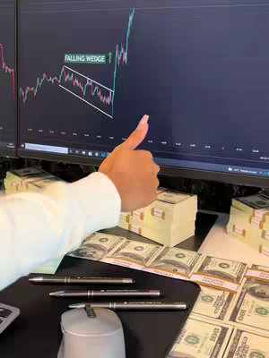 A person's arm with a thumbs-up gesture rests in the foreground. Behind it, two computer screens show a stock chart analysis including a 'Falling Wedge' pattern. Stacks of hundred-dollar bills, pens, and a mouse are visible on the desk. The composition suggests wealth and financial success, tying in with the business theme.