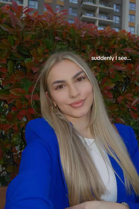 A close-up shot of a woman with long blonde hair, wearing a blue blazer over a white top. She is smiling slightly at the camera. Behind her is a bush with red and green leaves, and a building is visible in the background. The text "suddenly I see..." is overlaid on the image.