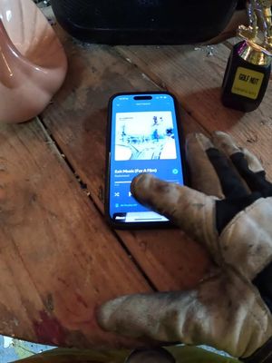 An image shows a close-up of a smartphone displaying a music app. The phone is on a wooden surface with a gloved hand touching the screen. In the background, there's a light-colored ceramic piece and a small bottle marked "GOLF NOT."
