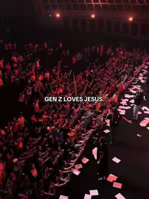 An overhead shot of a large gathering in an auditorium or similar space. A large crowd of people is visible, many with their arms raised, illuminated by red and pink lighting. Confetti or paper is scattered on the ground. The text 'GEN Z LOVES JESUS' is overlaid on the image.