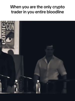 A grayscale image featuring two men at a conference or event. Text overlayed reads "When you are the only crypto trader in you entire bloodline." The background is dark, with a sign visible on the left reading "COMIC CON" and the Comic Con logo. The man on the right is in focus, wearing a light-colored button-down shirt.