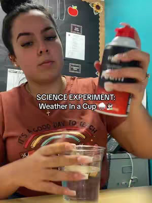 A woman is holding a can of whipped cream and a clear plastic cup filled with water. Text overlaid on the image states "SCIENCE EXPERIMENT: Weather In a Cup". The scene appears to be in a classroom setting.