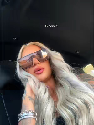 A woman with long, wavy, light-colored hair is the central focus, wearing large, dark sunglasses that reflect an outdoor scene. Her lips are prominently styled with a glossy finish. A silver bracelet adorns her wrist, and tattoos peek from beneath her sleeve. The background is darkly lit, possibly inside a vehicle, with overlaid text.