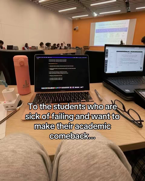 A medium shot shows a person's legs in gray sweatpants resting on a wooden desk with two open laptops and other items. The background includes a classroom setting with other students and a projection screen.