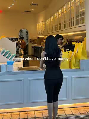 A woman in a black outfit stands with her back to the camera inside a cafe. Text overlay says 'me when i don't have money'. The background shows the interior of a cafe with windows reflecting sunlight.