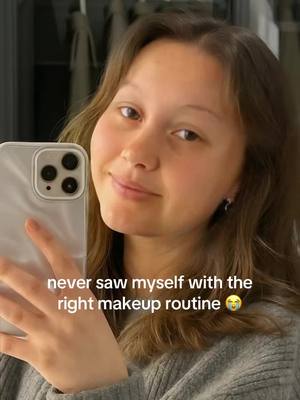 A selfie-style image featuring a young woman holding up a smartphone to capture the shot. She is smiling and looking at the camera, with a soft, natural expression. The background is simple, with a suggestion of a mirror or reflective surface behind her. The overall composition has a warm tone, emphasizing the subject.