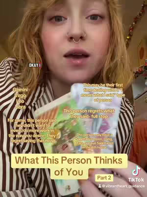 A woman with blonde hair is centered in the frame, looking towards the camera. She has a nose ring and is wearing a striped shirt. She is holding a tarot card and there are other tarot cards with text overlays. The overall composition is warm, the background is blurred.