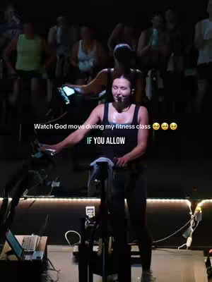 The image shows a woman on a stationary bike in a fitness class. She is positioned prominently in the center, with other individuals blurred in the background, also on bikes. The lighting is dim, suggesting a studio setting or a workout taking place during the evening. The text overlays emphasize the worship component of the class.