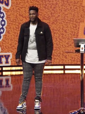 A man stands center-frame on a stage with an orange textured background, wearing a casual outfit: a black jacket over a white t-shirt, plaid pants, and colorful sneakers. The lighting suggests a stage setting, and the man appears to be the focus.