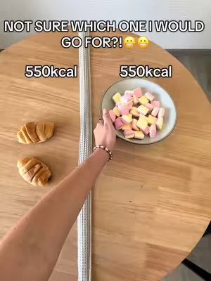 A top-down shot of a round wooden table. On the left, two croissants are placed. In the center, a long, gray textured strip runs vertically. On the right, a white bowl holds a pile of pink and yellow marshmallows, and a hand with a bracelet reaches towards the bowl.