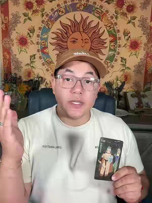 A man wearing glasses and a brown cap is holding a tarot card and speaking. The background consists of a tapestry with a sun design and floral patterns.