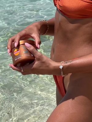 The cover image shows a person standing waist-up in the water, holding a brown jar, possibly skincare products, in both hands. The person is wearing a bright orange bikini and several gold bracelets. The background shows the clear blue water with some sunlight reflecting off the surface.