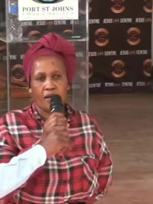 The image captures a medium shot of a woman speaking into a microphone. She is wearing a red and black plaid shirt and a red headscarf. Behind her is a banner displaying text, and the background has a patterned design repeating the words 'JESUS LIFE CENTRE'. The composition is straightforward.