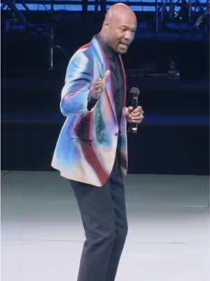 The image features a man on stage, posed as if mid-sentence. He's wearing a brightly colored, abstract-patterned jacket over a black shirt and pants. He holds a microphone, and the background is dark with hints of stage lighting. The overall composition emphasizes his expressive gesture, the colors of his clothes, and the setting of the stage.